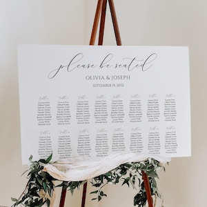 May include: A white rectangular wedding seating chart sign with black text reading please be seated, OLIVIA & JOSEPH, SEPTEMBER 15, 2032, and guest names. The sign is displayed on a brown wooden tripod easel decorated with green foliage and fabric.