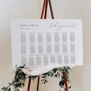 May include: A white rectangular wedding seating chart sign with black text reading Jessica & Brian, April 17, 2032, and take your seat, listing guests for tables 1 through 22. The sign rests on a brown wooden easel decorated with green foliage.