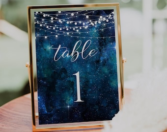 May include: A gold-framed table number card with a dark blue watercolor galaxy background and white string lights. The number "1" is written in white on the card. The card says "table" in white cursive writing.