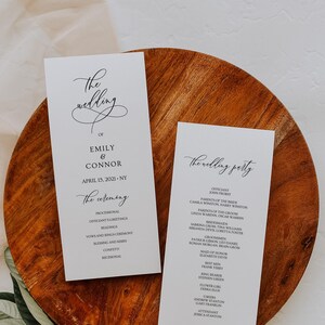 Op de afbeelding: Twee huwelijksceremonieprogramma's op een houten dienblad met groen. De programma's zijn wit met zwarte tekst, waaronder de namen "Emily & Connor" en de datum "15 april 2021". Eén programma bevat de ceremoniedetails, de andere de bruiloftsgasten.