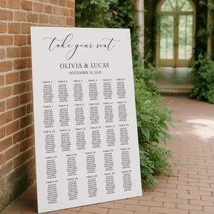May include: A white rectangular wedding seating chart sign with black text. The header reads take your seat, followed by OLIVIA & LUCAS and SEPTEMBER 19, 2028. The sign lists guest names organised under twenty-eight numbered table headings in columns.