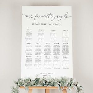 May include: A white wedding seating chart with the text "our favorite people PLEASE FIND YOUR TABLE". The chart lists names under table numbers 1-14. The names are in black font. The names are for Emily & Lucas, dated 08.09.2019. The chart is on a wooden easel.