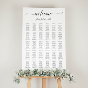 May include: A white seating chart with black text listing table numbers and guest names. The chart is on an easel with greenery in front of it. The text at the top reads "welcome please find your table".