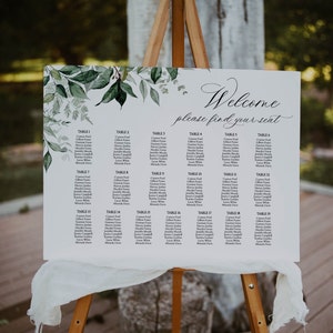 Puede incluir: Un diagrama de asientos de boda blanco con follaje verde y el texto "Bienvenido, por favor, encuentre su asiento". El diagrama enumera los nombres de los invitados y los números de mesa.