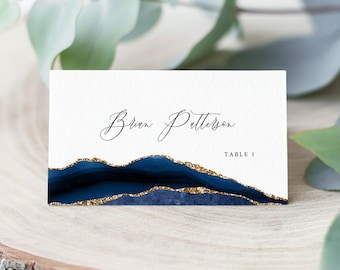May include: White place card with a blue and gold abstract design. The text "Brian Patterson" is written in black cursive script. The text "Table 1" is written in black sans-serif font.