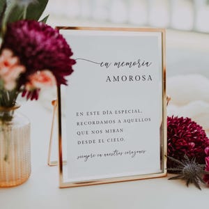 May include: A gold-framed sign with the words "en memoria AMOROSA" in elegant script. The sign includes additional text in Spanish, surrounded by floral arrangements in shades of pink and burgundy.