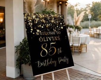 Può includere: Un cartello nero su un cavalletto di legno con testo dorato che recita "Benvenuti alla festa per l'85° compleanno di Olivia". La parte superiore del cartello presenta un design glitterato dorato. Lo sfondo mostra un evento all'aperto.