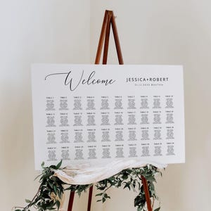 Peut inclure: Un panneau de plan de table de mariage rectangulaire blanc sur un chevalet en bois. Le panneau affiche le texte Welcome, Jessica + Robert, 11.11.2032 Boston, et une liste de noms d'invités organisée par numéros de table de 1 à 40.