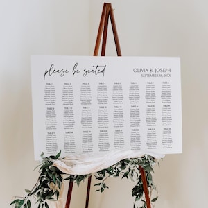 Könnte beinhalten: Ein weißes, rechteckiges Sitzplan-Schild für Hochzeiten auf einer Holzstaffelei mit dem Text please be seated, OLIVIA & JOSEPH, SEPTEMBER 15, 20XX, sowie einer Liste der Tische 1 bis 24 mit Namen, verziert mit einem weißen Stofftuch und grünen Blättern.