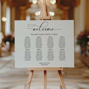 May include: A white wedding seating plan on a wooden easel, with names listed under table numbers. The sign has elegant calligraphy and the words 'welcome' and 'Please Find Your Table'. The wedding date is displayed.