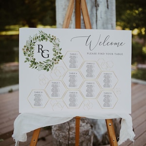 May include: A white wedding seating chart with a green and gold floral wreath featuring the initials "R" and "G" at the top. The chart is divided into hexagonal sections, each labelled with a table number and a list of guest names. The text "Welcome" and "Please find your table" is at the top of the chart.