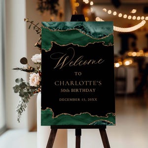 May include: A welcome sign for a 50th birthday celebration. The sign features a black background with emerald green and gold accents. The text reads "Welcome to Charlotte's 50th Birthday, December 15, 20XX."