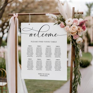 May include: A white rectangular wedding seating chart sign with black text reading "welcome" and "PLEASE FIND YOUR TABLE" followed by eight table lists and the names "EMILY & LUCAS 08.09.2032". The sign hangs from a wooden frame decorated with pink ribbons and a floral arrangement of white and pink roses.