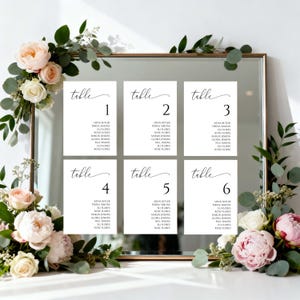 May include: Six white table cards with black calligraphy, each listing names, are arranged on a mirror. The cards are numbered 1 through 6. Floral arrangements with pink and white roses and greenery frame the mirror.