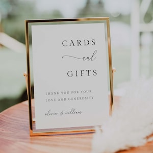 May include: A gold-framed sign with the words "CARDS and GIFTS" in elegant black script. Below, it reads "THANK YOU FOR YOUR LOVE AND GENEROSITY" followed by names. The sign is a wedding or event decoration.