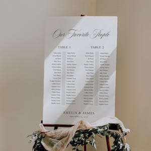以下が含まれることがあります： Our Favorite Peopleと題された白い長方形の結婚式用座席表。Table 1とTable 2のリスト、Amelia & Jamesの名前、September 15, 2022の日付が記載され、ベージュの布と緑の植物で装飾された金属製のイーゼルに立てかけられています。