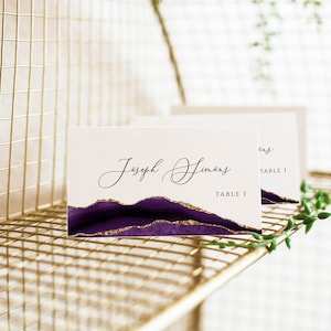 May include: Purple and gold wedding table card with the name "Joseph Simons" and the text "Table 1".