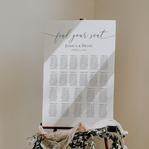May include: A white wedding seating chart sign on a wooden easel, featuring the text "find your seat", "JESSICA & BRIAN", and "APRIL 17, 2032", followed by a list of tables numbered 1 through 22 with names, decorated with draped beige fabric and greenery.