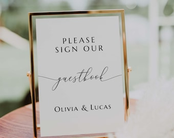 May include: A gold-framed sign with the text "PLEASE SIGN OUR guestbook" in elegant script. Below, it reads "OLIVIA & LUCAS". The sign is displayed on a wooden surface, with a blurred background.
