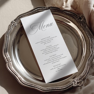 May include: A vertical white menu card with black serif and script typography featuring sections for appetizer, salad, entree, and dessert, resting on a polished silver charger plate with a decorative scalloped edge and a textured surface.