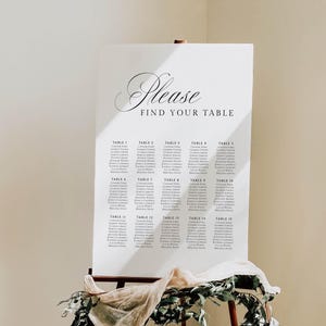 Puede incluir: Un cartel blanco de distribución de mesas para bodas sobre un caballete de madera, con el texto "Please FIND YOUR TABLE" en negro, seguido de quince mesas con nombres de invitados, decorado con tela beige y follaje verde.