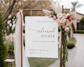 May include: A white sign with the words "WELCOME TO OUR rehearsal DINNER" in black script. The sign is part of a wooden frame decorated with flowers and greenery. The date "September 17, 2026" is also printed on the sign.
