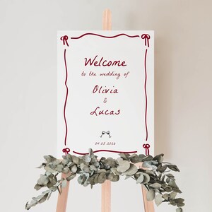 May include: White wedding welcome sign with red border and text that reads "Welcome to the wedding of Olivia & Lucas 04.05.2026" with a small image of two wine glasses.