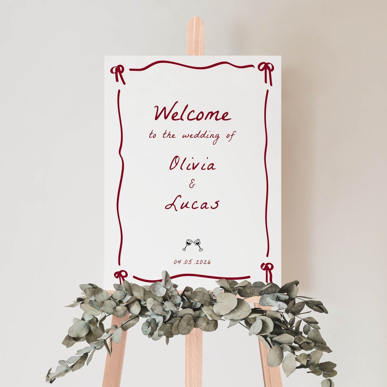 May include: White wedding welcome sign with red border and text that reads "Welcome to the wedding of Olivia & Lucas 04.05.2026" with a small image of two wine glasses.