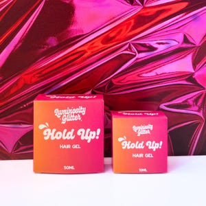 Puede incluir: Dos cajas de gel para el cabello "Luminosity Glitter Hold Up!". Las cajas son rosas y naranjas con texto blanco. La caja más grande contiene 50 ml de producto, y la caja más pequeña contiene 10 ml. El fondo es una superficie rosa brillante y texturizada.