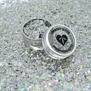 May include: A silver glitter in a small tin container. The container has a black label that reads "Luminosity Glitter Biodegradable Glitter".