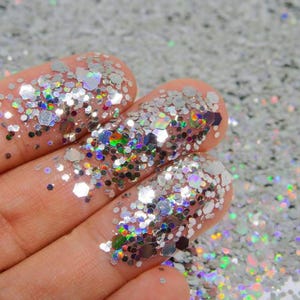 May include: A collection of loose silver glitter particles featuring a mix of hexagonal and fine shapes. The glitter reflects light with a holographic, multi-colored sheen, creating a bright, metallic, and shimmering effect across the surface.
