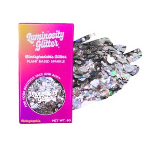 Op de afbeelding: Een roze doos met holografische Luminosity Glitter voor gezicht en lichaam. De doos heeft de tekst "Luminosity Glitter" in witte letters en "Biodegradable Glitter, Plant Based Sparkle". Naast de doos ligt een hoop zilveren glitter. Nettogewicht: 6g.