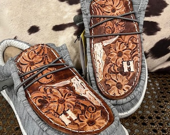 May include: A pair of gray slip-on shoes with brown leather accents. The leather features a floral design and the letter "H". The shoes have gray laces and white soles. The background is a brown and white cowhide pattern.