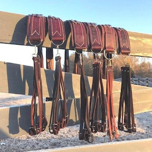 May include: A row of leather horse tack, including headstalls and reins, hangs on a wooden fence. The leather is a rich brown color with white stitching. The metal hardware is silver. The word "OPEN" is visible on some of the tack.