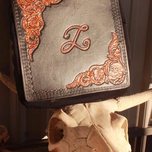 May include: A square black leather patch featuring a central embossed letter L, surrounded by intricate floral and skull tooling in a natural tan finish. The border is detailed with a stamped geometric pattern, creating a textured contrast.