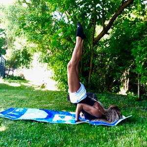 May include: A person doing a handstand on a blue and white yoga mat in a grassy area. The person is wearing black socks, white shorts, and a black top. Green trees are in the background.