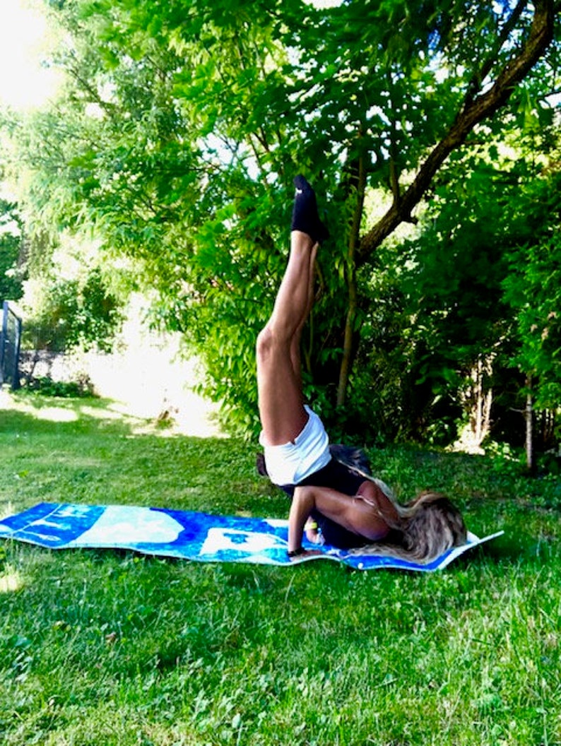 May include: A person doing a handstand on a blue and white yoga mat in a grassy area. The person is wearing black socks, white shorts, and a black top. Green trees are in the background.