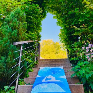 May include: A blue yoga mat with white yoga poses is laid out on a concrete staircase leading to a bright, sunny opening. The mat has the text "SUNRISE YOGA" at the bottom. The staircase is surrounded by lush green foliage.