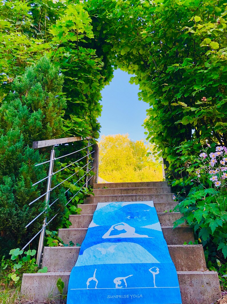 May include: A blue yoga mat with white yoga poses is laid out on a concrete staircase leading to a bright, sunny opening. The mat has the text "SUNRISE YOGA" at the bottom. The staircase is surrounded by lush green foliage.