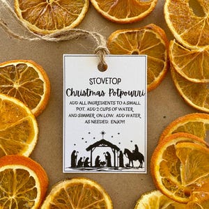 Könnte beinhalten: Ein weißes Etikett mit dem Text "STOVETOP Christmas Potpourri" und Anweisungen, umgeben von getrockneten Orangenscheiben. Das Etikett zeigt eine schwarze Silhouette der Krippenszene. Die Anweisungen sind in Schwarz gedruckt.