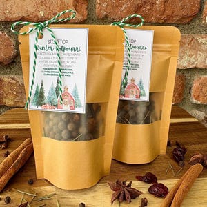 May include: Two kraft paper pouches of stovetop potpourri with a clear window, tied with green and white twine. Each pouch has a label with instructions and a winter scene illustration. Cinnamon sticks, star anise, and cranberries are scattered around.