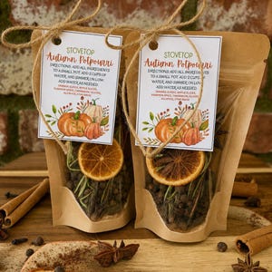 May include: Two brown paper pouches containing stovetop autumn potpourri, each tied with twine and a white tag. The tag features text, pumpkin illustrations, and lists ingredients like orange slices, apple slices, pumpkin spice, cinnamon, cloves, allspice, and star anise. Dried orange slices are visible through the clear plastic window of each pouch.