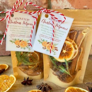 May include: Two clear pouches filled with dried orange slices, cinnamon sticks, and rosemary, labeled "Stovetop Christmas Potpourri." Each pouch is tied with red and white twine and includes a tag with instructions and ingredients.