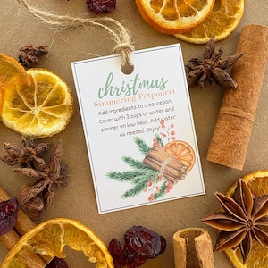 Op de afbeelding: Een label met het woord "christmas" en instructies voor het maken van sudderpotpourri. Het label is vastgebonden met touw en omgeven door gedroogde sinaasappelschijfjes, kaneelstokjes, steranijs en veenbessen.