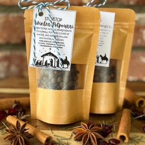 May include: Two brown paper pouches of stovetop winter potpourri with a clear window, tied with blue and white twine. The label reads "Stovetop Winter Potpourri" with instructions and a nativity scene. Cinnamon sticks, star anise, and cranberries are scattered around.