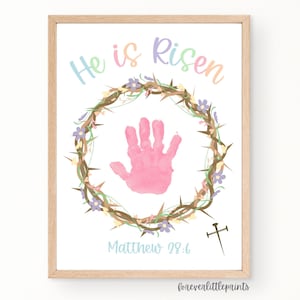May include: A framed print with the words "He is Risen" in pastel colors above a pink handprint inside a wreath of thorns and flowers. The text "Matthew 28:6" is below the handprint, with a cross to the right. The frame is light brown.