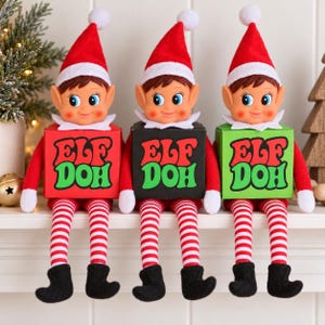 Peut inclure: Trois poupées lutins portant des bonnets rouges avec des pompons blancs, des hauts rouges à manches longues et des leggings rayés rouge et blanc avec des bottes noires. Chaque lutin tient une boîte carrée marquée ELF DOH en rouge et vert.