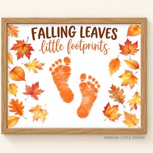 May include: A framed print featuring the text FALLING LEAVES little footprints in brown and orange script. The design includes orange baby footprints in the center surrounded by watercolor illustrations of autumn leaves in shades of red, orange, and yellow.