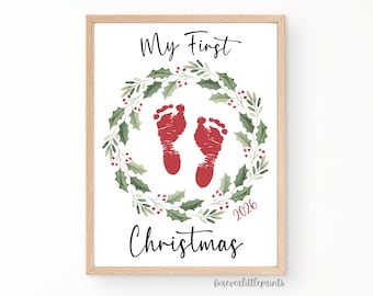 May include: A framed print featuring the text My First Christmas in black script, surrounding a watercolor wreath of green holly leaves and red berries. In the center are two red baby footprints, with the year 2026 printed in red below.