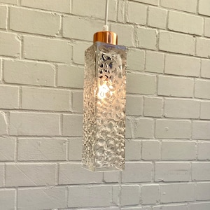 May include: A clear, rectangular pendant light with a textured glass shade. The light fixture has a copper-coloured top and a white cord. The light is illuminated, casting a warm glow. The light hangs in front of a white brick wall.